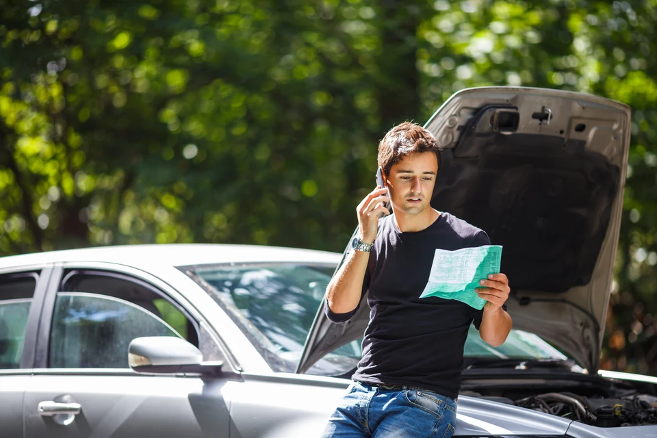 Homme utilisant un téléphone portable près d'une voiture avec le capot ouvert, tenant des documents dans un environnement extérieur arboré