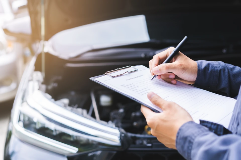 Extérieur automobile, professionnel en chemise bleue complétant un formulaire sur presse-papiers près d'un véhicule avec capot ouvert
