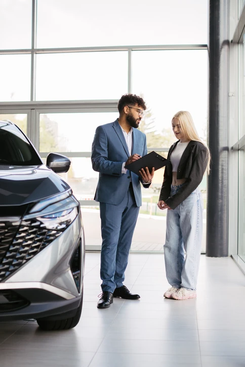 Femme en pull gris examinant une petite voiture bleue miniature avec une loupe dans un intérieur moderne avec canapé bleu en arrière-plan